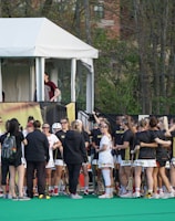 A group of athletes in sports uniforms is gathered on what appears to be a lacrosse field. They are standing in two groups, some wearing white uniforms and some wearing dark uniforms. There is a tent with people inside, possibly officials or commentators, overlooking the field in the background. Extensive greenery and trees are visible behind the tent.